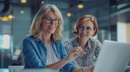 
Two happy busy female employees working together using computer planning project. Middle aged professional business woman consulting teaching young employee looking at laptop sitting at desk in offic