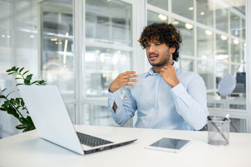 A young businessman loosens his tie, feeling hot and stressed in a bright, modern office setting. The heat seems too much as he cools himself with his hand.