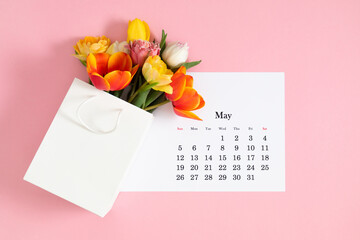 Flat lay of paper desk calendar for May 2024, top view. A bouquet of bright colorful tulips on pastel pink background