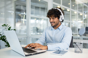 Obraz premium A cheerful young man with curly hair wears headphones while focusing on work using a laptop in a modern office environment, showcasing productivity and concentration.