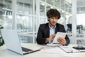 A focused young businessman analyzes data on a tablet while sitting at a desk in a bright, contemporary office. Essential tools and documents surround him.