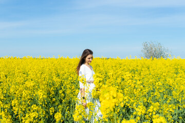 Young beautiful woman in a white dress close up in the middle of the yellow field with the radish flowers closeup on the blue sky background. Republic of Belarus, Brest region