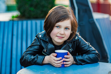 A girl with a square hairstyle, sitting in a summer cafe and drinking tea from a disposable cup, with a guitar in the background