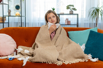 Ill girl child suffering from cold flu allergy infection wrapped in blanket sitting on sofa in living room at home. Sick teenager blows sneezes wipes snot into napkin. Coronavirus quarantine pandemic