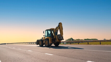 Yellow backhoe loader traveling on highway under clear blue sky signifying ongoing construction...