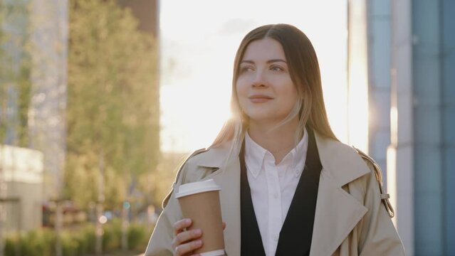 Stylish young woman in a coat walking during sunset around the city holding a cup of coffee 