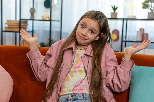 I don't know what to say. Confused young girl child feeling embarrassed about ambiguous question, having doubts, no idea, being clueless and uncertain. Teen kid sitting on couch in living room at home