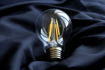 Modern clear LED filament lamp with yellow filaments on dark silk fabric. Photo. Copy space