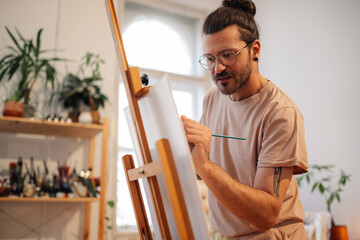 Focused hipster artist with brush painting on easel at creative atelier