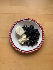 A plate with cottage cheese, sour cream and blueberries on a round tray in a white and red small checkered pattern stands on a wooden background. View from above. Place for text