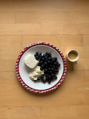 A cup of espresso coffee and a plate with cottage cheese, sour cream and blueberries on a round tray in a white and red small checkered pattern stand on a wooden background. View from above. 