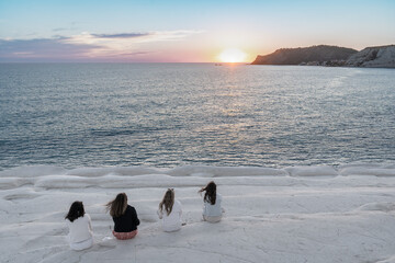 quatre jeunes filles de dos assises sur des rocher blancs face à un coucher de soleil sur la mer