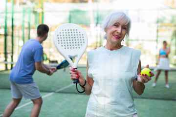 Portrait of positive elderly woman standing on padel tennis court, holding racket and ball and...