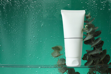 Tube with moisturizing cream and eucalyptus branches on green background, view through wet glass....