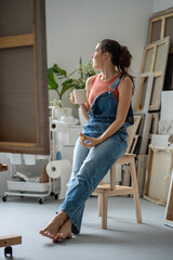 Artist lunch in home studio, freelancer working break. Young female painter relaxes with coffee cup, small snack after work. Barefoot woman with paintings eating at workplace, creativity concept - obrazy, fototapety, plakaty