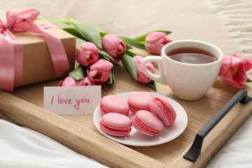 Tasty breakfast served in bed. Delicious macarons, tea, flowers, gift box and card with phrase I Love You on tray