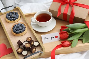 Tasty breakfast served in bed. Delicious desserts, tea, flowers, gift box and card with phrase I Love You on tray