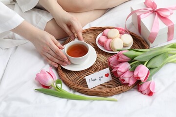 Tasty breakfast served in bed. Woman with tea, macarons, gift box, flowers and I Love You card at home, closeup
