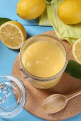 Delicious lemon curd in glass jar, fresh citrus fruits, green leaves and spoon on light blue wooden table
