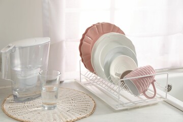 Drainer with different clean dishware, cup, glass and filter jug on light table indoors