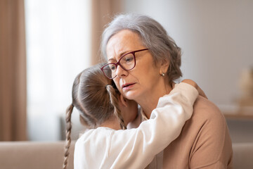 A grandmother with glasses and a caring expression is hugging her young granddaughter, who seems to...