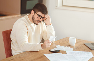A man is seated at table, concentrating on papers in his hand with a thoughtful expression. Scattered documents, a calculator on the table, indicating a session of financial review or budgeting