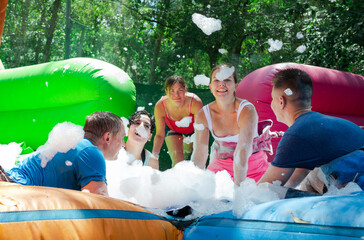 Cheerful young adult friends having fun in outdoor amusement park on sunny summer day, picking up balls in inflatable pool full of foam..