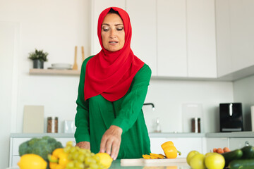 A woman wearing a red and green hijab is shown in the act of chopping a variety of vegetables on a cutting board.