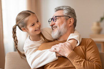 Senior man and a little girl are seated on a couch, looking towards each other, grandfather and granddaughter embracing at home