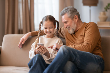 A smiling grandfather is sitting closely to his young granddaughter on a beige sofa, sharing a...