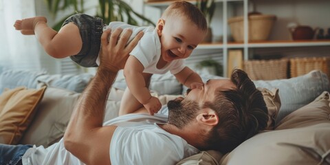 A handsome young father is lying on the sofa, holding his one-year-old baby boy in midair and lifting him up with both hands to play