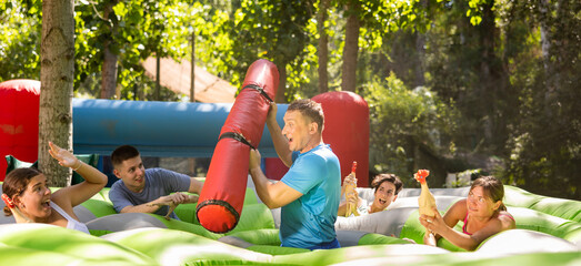 Happy playful man playing with friends in amusement park on summer day, fighting back with inflatable log while guarding toy chickens..