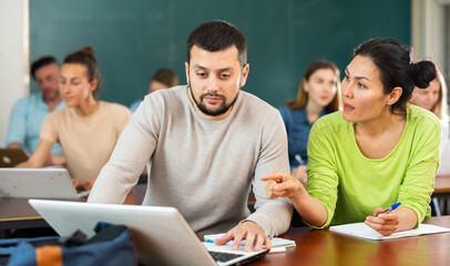 Asian woman and european man university students sitting at table in classroom, using laptop and...