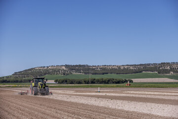 Fototapeta premium A green tractor pulling a red plow through a crop field with a forested mountain behind
