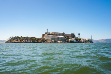 Alcatraz Island view under clear sunny skies, featuring Alcatraz Federal Penitentiary structures from the bay in San Francisco Bay. Notable landmarks visible.