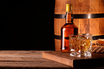 Whiskey with ice cubes in glass, bottle and barrel on wooden table against black background, space...
