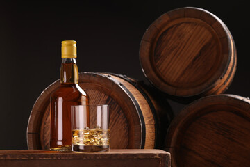 Whiskey with ice cubes in glass and bottle on wooden table near barrels against black background,...