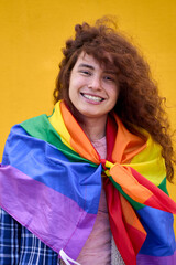 Vertical. Portrait of cheerful non-binary person with red hair with rainbow flag smiling looking at camera on yellow background. LGBT community, diversity, transgender and non-binary people. 