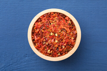 Chili pepper flakes in bowl on blue wooden table, top view