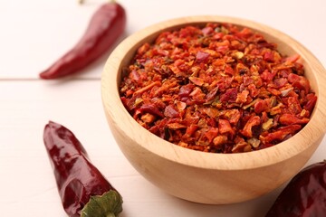 Chili pepper flakes in bowl and pods on white wooden table, closeup