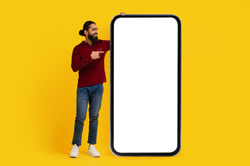 Indian man with a beard is cheerfully pointing at a giant, blank smartphone screen while standing...