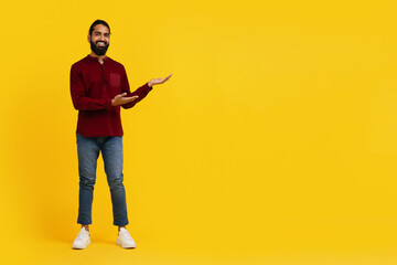 A cheerful Indian man stands against a vivid yellow backdrop, looking at the camera with a confident smile, gesturing with his hands open, copy space