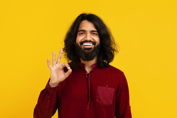 Indian man with a full beard and long hair smiles broadly, standing against a vivid yellow...