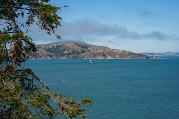 Serene body of water with sailboat, coniferous tree branches, hilly landscape, clear sky in...