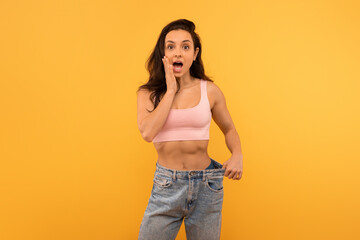 A young woman stands with a shocked expression on her face, her hand touching her cheek, wearing a casual pink crop top paired with oversized blue denim jeans