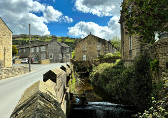 Holme Beck flows beside a stone wall and beneath a charming road, with ancient stone cottages and green hills in the tranquil rural village of Sutton-in-Craven, Yorkshire, UK.