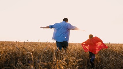 Father and son superhero in red cloak flying running at sunny dry wheat field back view. Happy...