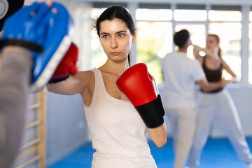 Sportive young female practitioner of boxing courses applying kicks on hitting mitts during workout...