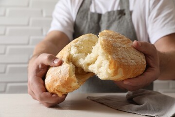 Man breaking loaf of fresh bread at white table near brick wall, closeup