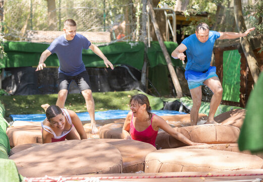 Group of cheerful young adults having fun in green summer amusement park, moving through obstacle course with soft pillows in pool of water..